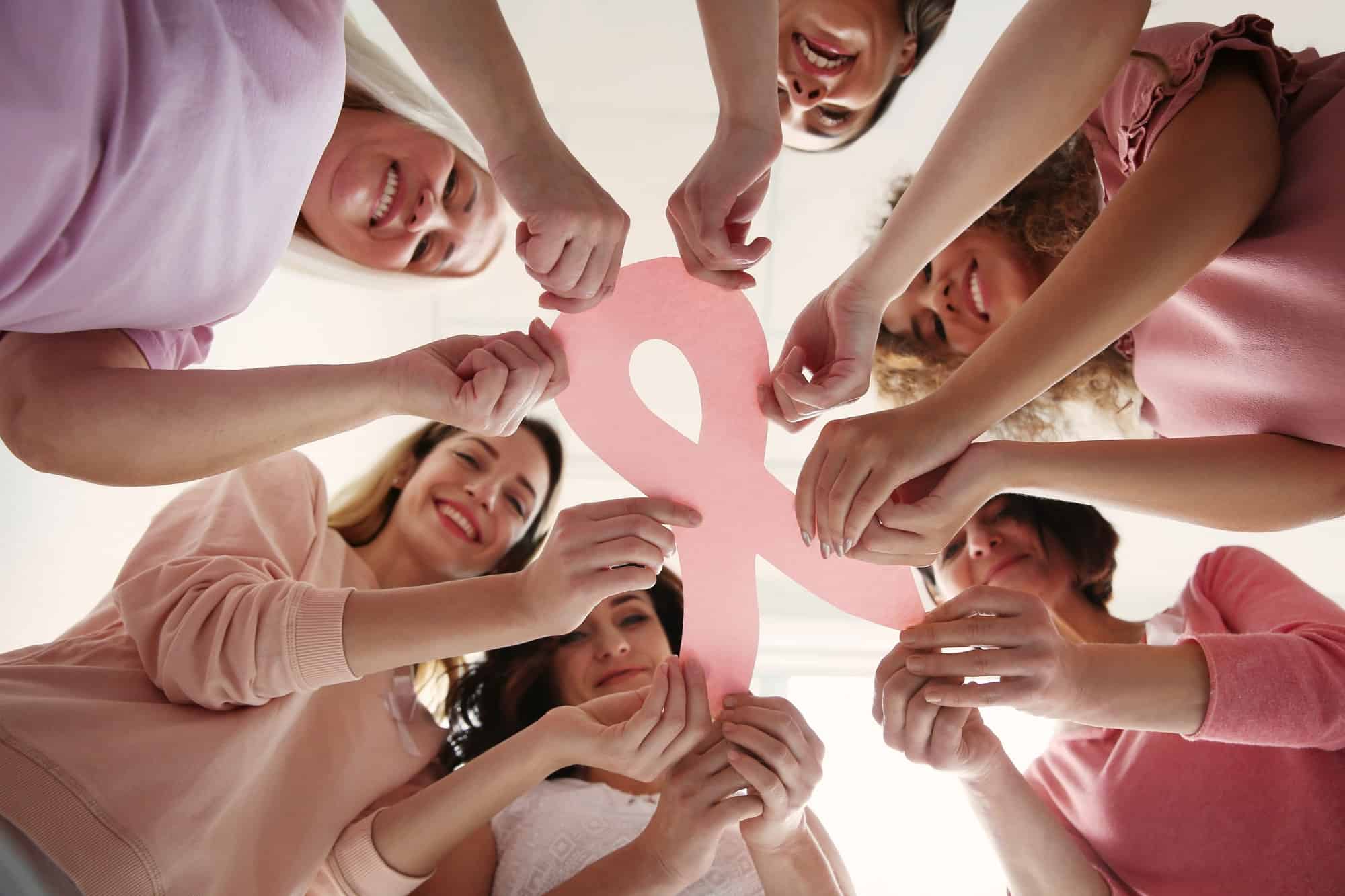 group of women wearing pink shirts holding pink paper cutout of ribbon for breast cancer awareness