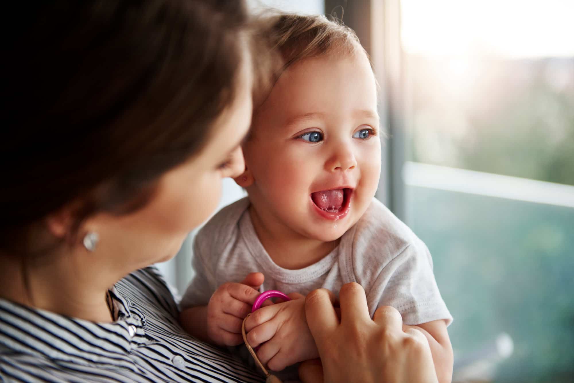 woman holding small baby with mouth open smiling near window