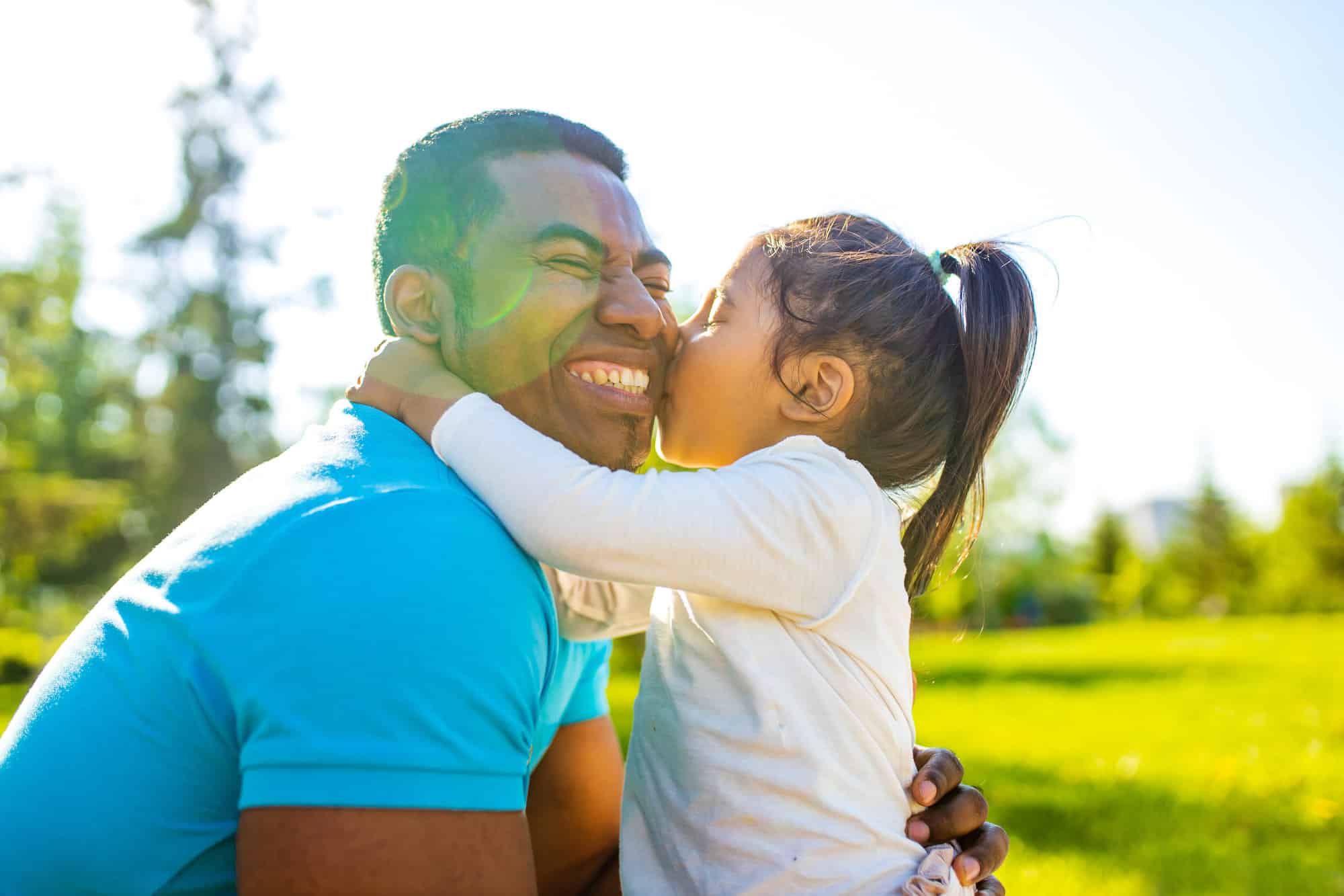 young girl kissing smiling father on cheek sunny outdoors