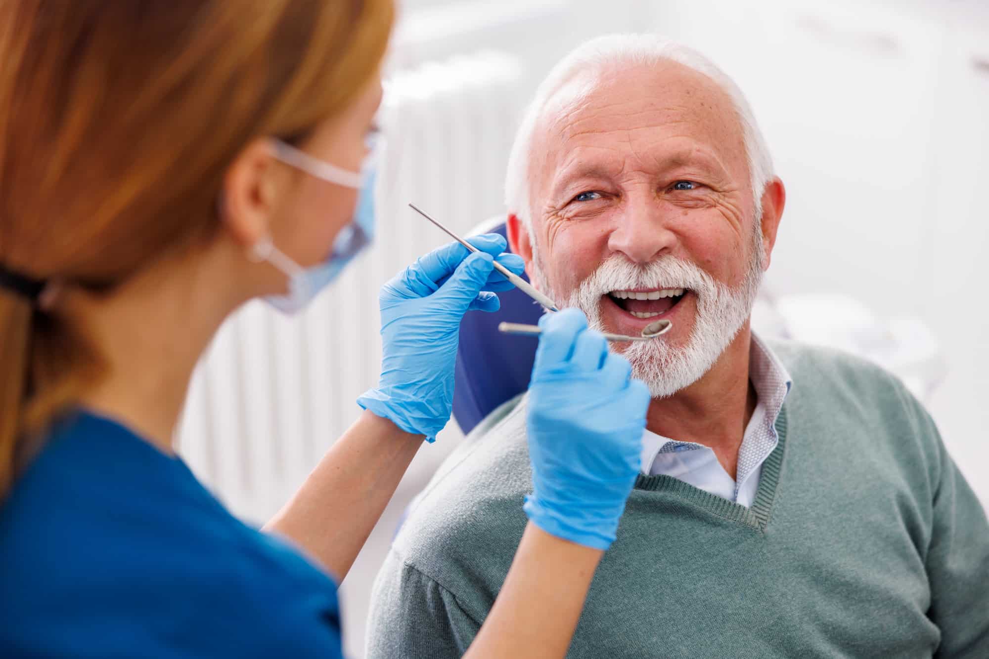 older man smiling getting teeth checked by dental assistant holding metal tools