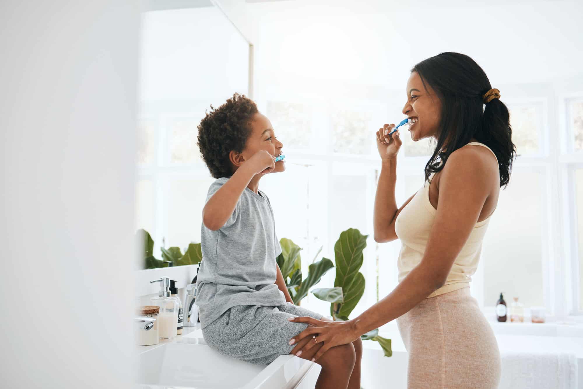 young child sitting on the counter brushing teeth with mom in light bathroom