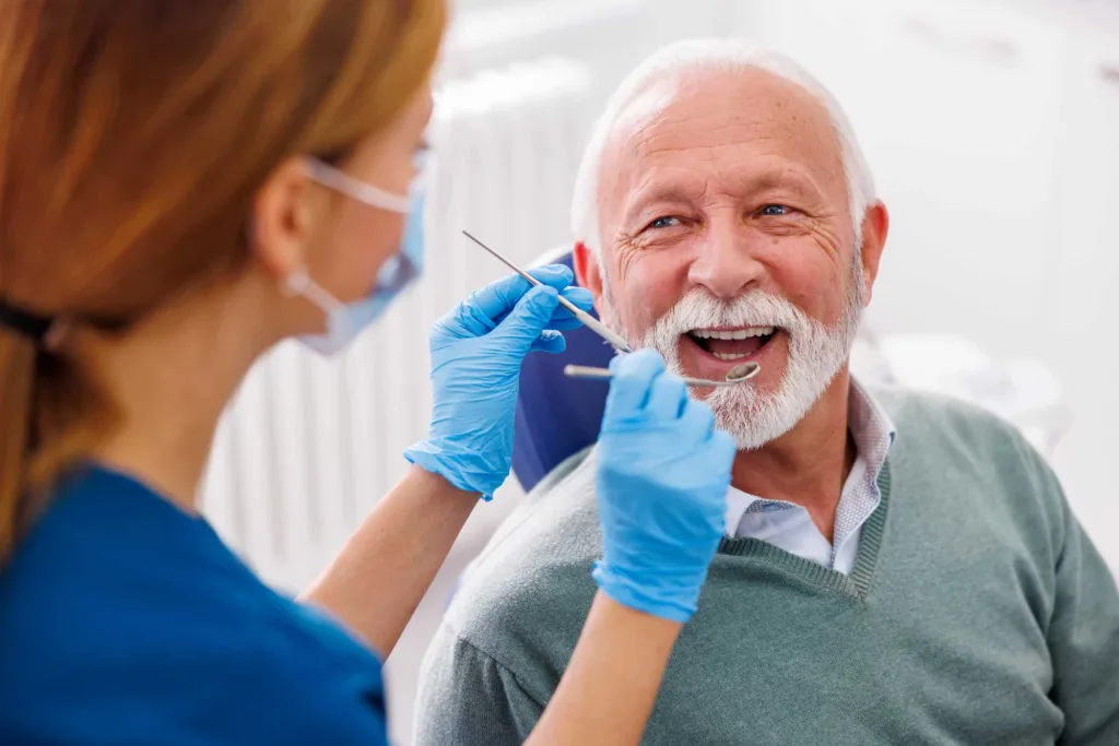 older man smiling getting teeth checked by dental assistant holding metal tools